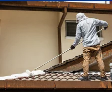 A person wearing protective clothing and a hat is standing on a tiled roof in Georgia, using a long-handled roller to apply white paint or sealant—showcasing the expert Roofing Services offered by Prime Roofing and Restoration.