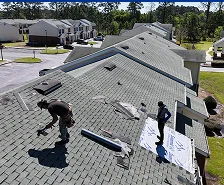 Two workers from Prime Roofing and Restoration are repairing a residential roof in Georgia; one uses a tool while the other stands on roofing underlayment. Several buildings, trees, and a parking lot are visible in the background.