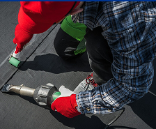 A person wearing red gloves and a plaid shirt uses a heat welding tool to seal black roofing material, showcasing the expert work of Prime Roofing and Restoration, trusted for quality roofing services across Georgia.