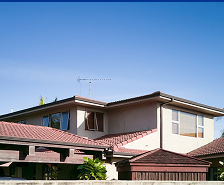 A two-story house with brown tiled roofs by Prime Roofing and Restoration, beige walls, and large windows, surrounded by greenery under a clear blue Georgia sky.
