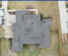 Aerial view of a large house with a gray, multi-peaked roof in Georgia, surrounded by a patchy lawn and driveway. Prime Roofing and Restoration offers expert roofing services for homes like this one.